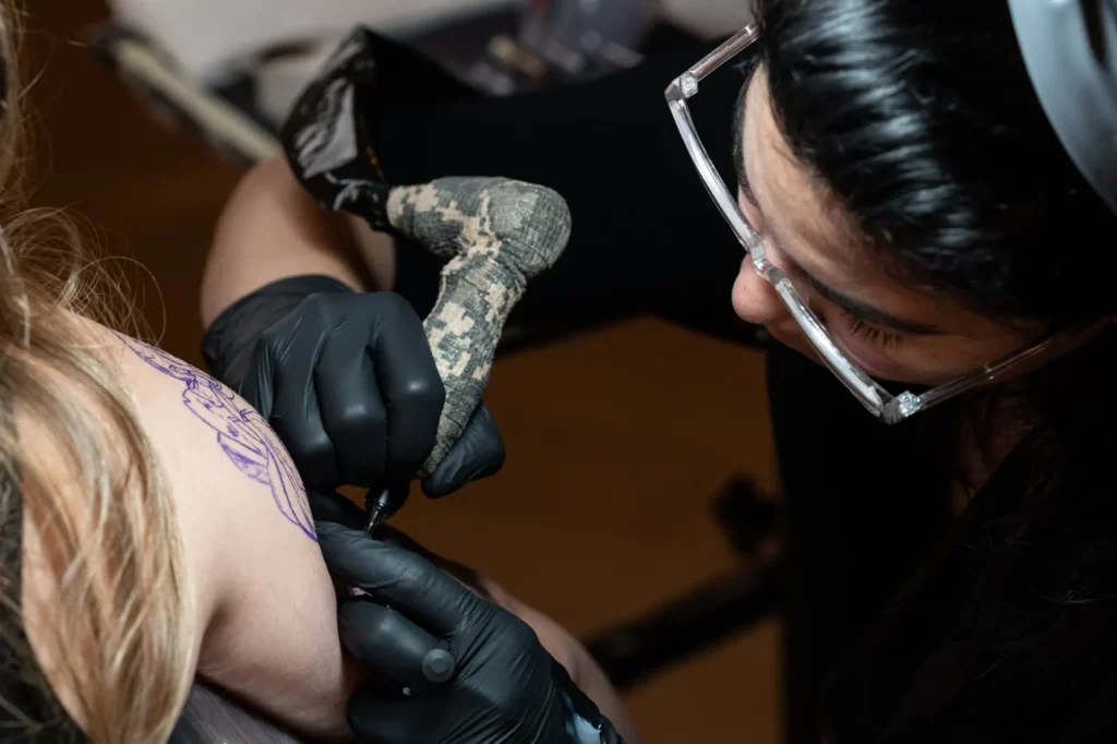 Tatuador con guantes negros y gafas de protección tatuando un hombro con el diseño marcado en la piel.
