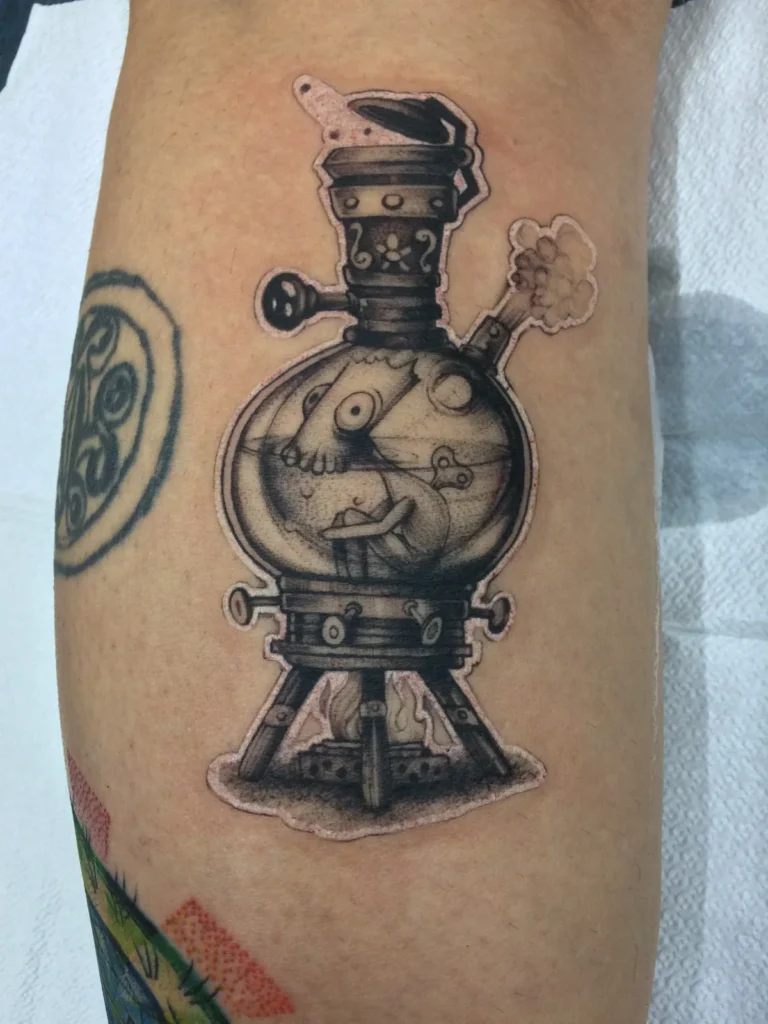 Tatuaje en blanco y negro de un alambique ilustrado con un personaje dentro, en la pierna, hecho en Ochoa Ink