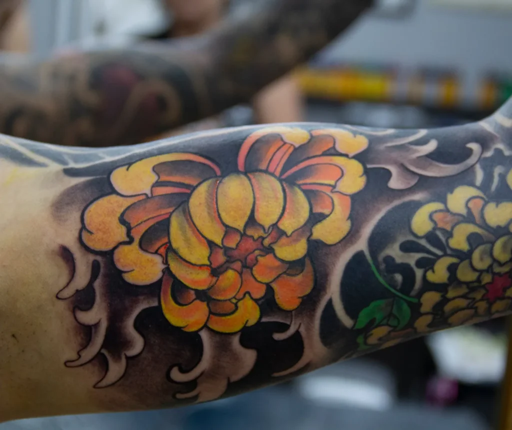Tatuaje neoriental japonés a color de un crisantemo amarillo y naranja entre olas, en el brazo